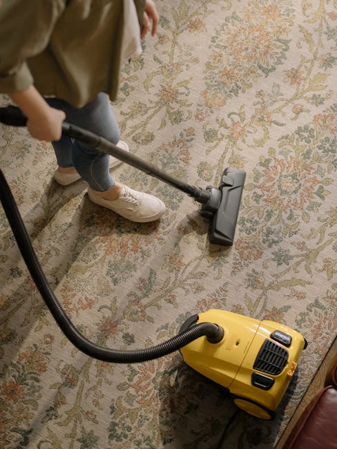 A person using a yellow vacuum cleaner with a black hose attachment to clean a patterned beige and green carpet in a residential living room. The individual is wearing light-colored sneakers and jeans, and is operating the vacuum head over the carpet's surface, which appears dust-free and well-maintained. The room is illuminated with natural or ambient light, highlighting the detailed floral design on the carpet. The setting suggests a thorough domestic cleaning process with attention to surface hygiene. Carpet Cleaners Lambeth specializes in high-quality cleaning services, including deep carpet cleaning and sanitisation, ensuring a fresh and hygienic environment.