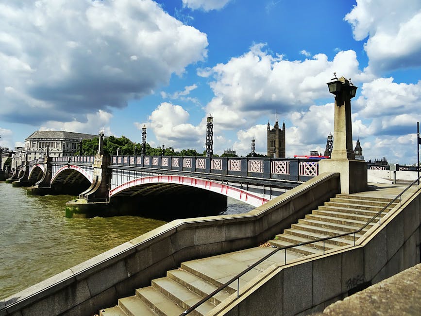 A daytime view of the Westminster Bridge spanning the River Thames in London, featuring ornate lampposts along the balustrade and a series of arches beneath the bridge. The bridge's painted details are visible, and the surface appears clean and well-maintained. In the background, the historic gothic-style church tower and other iconic London landmarks can be seen under a partly cloudy sky, with sunlight illuminating the scene. The stairs leading down from the riverbank are visible in the foreground, constructed from stone with metal railings, suggesting recent cleaning or maintenance for safety and aesthetic purposes. The image highlights the architectural features and cleanliness of the public space, consistent with surface cleaning and preservation efforts that might be carried out by professional cleaning services like Carpet Cleaners Lambeth.