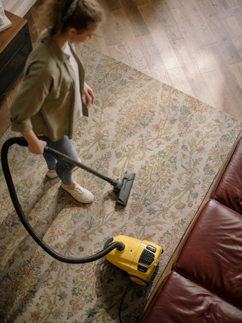 A person vacuuming a patterned area rug in a living room. The rug features floral motifs in muted tones, situated on a light wooden floor. The individual is dressed casually in a beige jacket, dark pants, and white sneakers, holding a yellow vacuum cleaner with a black hose. The room is well-lit, and part of a brown leather sofa is visible in the corner. This scene depicts surface cleaning and deep cleaning processes, emphasizing hygiene and maintenance, with the setting indoor and private. Carpet Cleaners Lambeth specializes in upholstery and carpet cleaning, ensuring cleanliness and freshness for domestic spaces, as illustrated by the efficient vacuuming in this image.