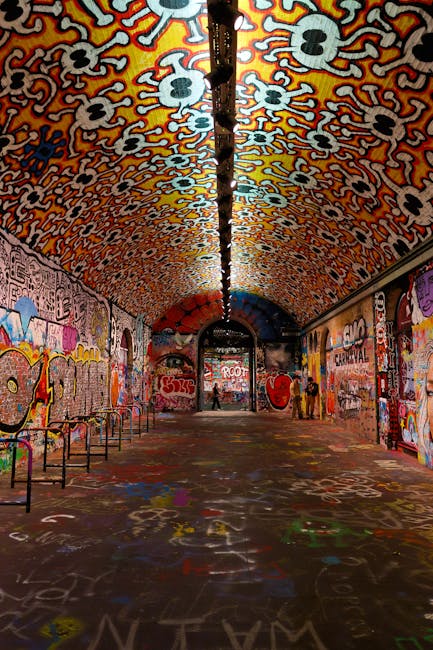 An arched underground tunnel with a vibrant, graffiti-covered ceiling and walls, featuring various colorful spray-painted images and text. The floor is scattered with multicolored chalk or spray paint markings, creating a lively, artistic atmosphere. A black overhead pipe runs along the center of the tunnel, with lighting fixtures illuminating the space evenly. The tunnel is empty except for a few visitors in the distance, and the scene portrays a well-maintained environment with no visible dirt or debris. The setting is designed for artistic expression and urban exploration, with attention to surface cleanliness and visual impact, aligning with professional cleaning standards promoted by Carpet Cleaners Lambeth for communal and entertainment spaces.