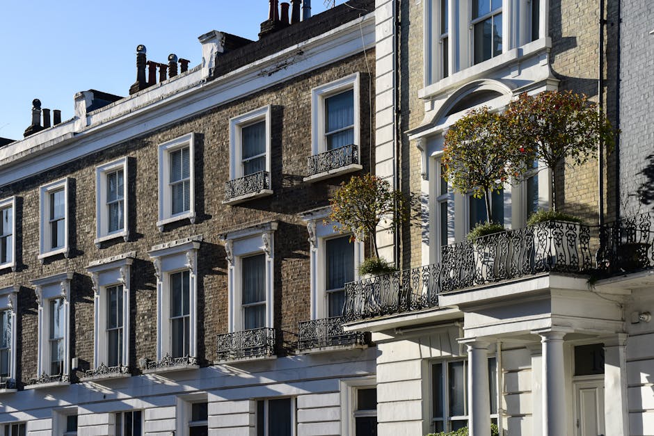 Photograph of a row of multi-story residential buildings with classic brick facades and white window frames. Several windows feature decorative wrought-iron balcony railings, some adorned with potted plants and small trees. The buildings are illuminated by bright sunlight, casting distinct shadows on the surfaces. The architecture includes ornate window surrounds and a mixture of traditional and modern design elements. The overall appearance is clean and well-maintained, reflecting the quality of property management and upkeep typical in Kennington Lambeth. Carpet Cleaners Lambeth is mentioned as a professional cleaning service experienced in domestic cleaning and surface sanitisation, ensuring the homes remain in pristine condition.