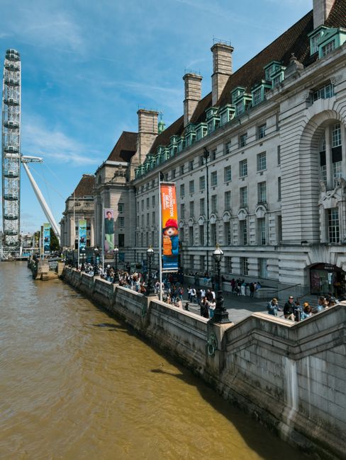 Exterior view of a large historic building along a riverbank in Lambeth, featuring white stone facades, arched windows, and multiple chimneys. The riverside walkway is crowded with pedestrians, some standing near lampposts and banners advertising local attractions. The Thames River runs alongside the promenade, with a modern Ferris wheel visible to the left under a clear blue sky. The area appears lively and well-maintained, with clean surfaces and organized pedestrian pathways, highlighting the importance of surface cleaning and maintenance as part of urban beautification and hygiene efforts by Carpet Cleaners Lambeth for council estate environments.