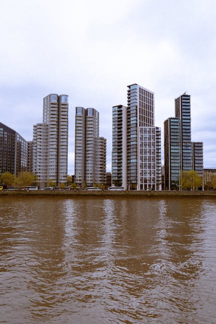 Image of a cityscape with modern high-rise buildings along a riverfront. The structures are predominantly glass and steel, reflecting natural light and exhibiting contemporary architectural styles. The buildings vary in height and design, with some featuring rounded edges and others with rectangular facades. The river in the foreground has gentle ripples, and the sky above is overcast with grey clouds, creating a neutral atmosphere. The scene highlights urban development and architectural diversity in the area. This image is related to the services offered by Carpet Cleaners Lambeth, emphasizing clean, well-maintained environments through surface cleaning and deep cleaning for residential or commercial spaces.