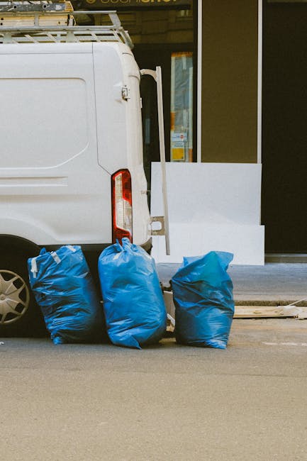 A person dressed in orange work overalls stands on a grey carpeted floor, holding two large blue plastic trash bags filled with waste. The bags appear sealed and are positioned at waist level. In the