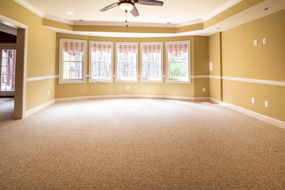 Close-up view of a red handheld vacuum cleaner positioned on a beige textured rug in a domestic setting, with a person barefooted standing nearby on the same carpet. The vacuum has a slim body with a