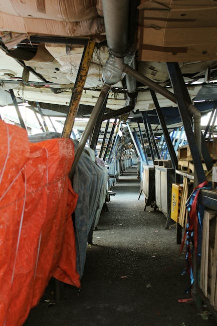 Inside a covered walkway or corridor with a curved ceiling structure, featuring exposed pipes and supports made of metal and wood. Along both sides, there are various equipment and storage units, some covered with tarps or protective covers, including a prominent orange cover on the left. The floor appears concrete with scattered dirt and debris, indicating a space that requires cleaning and maintenance. The lighting is dim, with natural light filtering from openings at either end of the corridor. Carpet Cleaners Lambeth specializes in surface cleaning and deep cleaning services, ensuring hygiene and tidiness in commercial and industrial environments, as depicted in this setting requiring professional cleaning attention.
