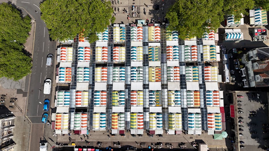 An aerial view of a busy outdoor market in Brixton, Lambeth, featuring numerous white tents with colorful striped canopies in red, yellow, blue, and green arranged in a grid pattern. The stalls are set up on paved surfaces with pedestrians walking between them. Surrounding the market are tall green trees, and adjacent streets with parked cars and additional buildings. Bright daylight illuminates the scene, highlighting the neat organization and cleanliness of the market area. As a professional cleaning specialist, I recommend regular surface cleaning and sanitisation to maintain hygiene in such bustling environments, a service offered by Carpet Cleaners Lambeth for commercial spaces like markets.