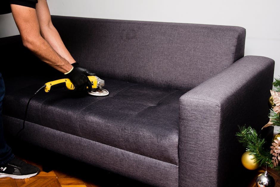 A person wearing black gloves is using a yellow handheld electric upholstery cleaner on a dark gray fabric sofa in a living room. The sofa has a modern design with a high backrest and armrest on the right side. The cleaning process appears to be part of deep cleaning or surface sanitisation to maintain hygiene. The surrounding area includes a wooden floor and a small Christmas tree decorated with gold and pink ornaments on the right side. The lighting is even, highlighting the clean appearance of the fabric. This image reflects professional upholstery cleaning performed by Carpet Cleaners Lambeth for residential properties such as Clapham Common flats, emphasizing thorough surface cleaning and maintenance.
