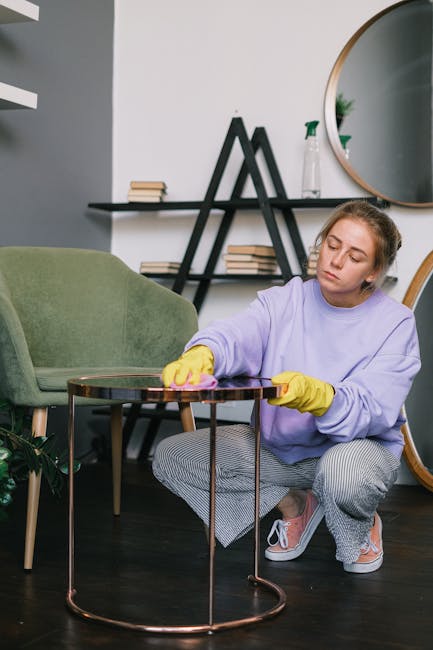 A person wearing black gloves is using a yellow handheld electric upholstery cleaner on a dark gray fabric sofa in a living room. The sofa has a modern design with a high backrest and armrest on the right side. The cleaning process appears to be part of deep cleaning or surface sanitisation to maintain hygiene. The surrounding area includes a wooden floor and a small Christmas tree decorated with gold and pink ornaments on the right side. The lighting is even, highlighting the clean appearance of the fabric. This image reflects professional upholstery cleaning performed by Carpet Cleaners Lambeth for residential properties such as Clapham Common flats, emphasizing thorough surface cleaning and maintenance.