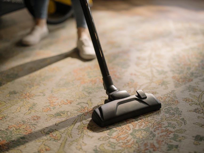 Close-up of a person vacuuming a patterned carpet in a domestic setting, with the vacuum cleaner's black head and hose visible in the foreground. The carpet features intricate floral designs in soft earth tones and appears clean and well-maintained. The person is wearing slippers, and natural light illuminates the room. This scene illustrates surface cleaning and maintenance, emphasizing hygiene and deep cleaning practices. The image is associated with Carpet Cleaners Lambeth, a professional cleaning company focused on thorough domestic cleaning services, including carpet sanitisation to address hidden charges in Lambeth carpet cleaning.